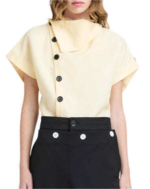 A woman stylishly wears the Proenza Schouler Zion Top, a beige asymmetrical buttoned piece with a cowl neck and short sleeves, paired with black pants accented by white buttons.