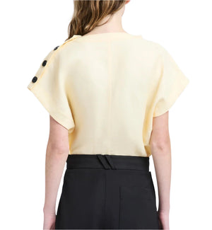 Seen from behind, the person wears a cream Zion Top by Proenza Schouler with black shoulder buttons, paired with high-waisted black pants. The top’s draped silhouette adds elegance to the look.
