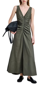 A woman wears the Remi Dress by Proenza Schouler—an olive green Eco Stretch Linen maxi with a deep V-neckline and gathered details—paired with a black bag and black loafers.