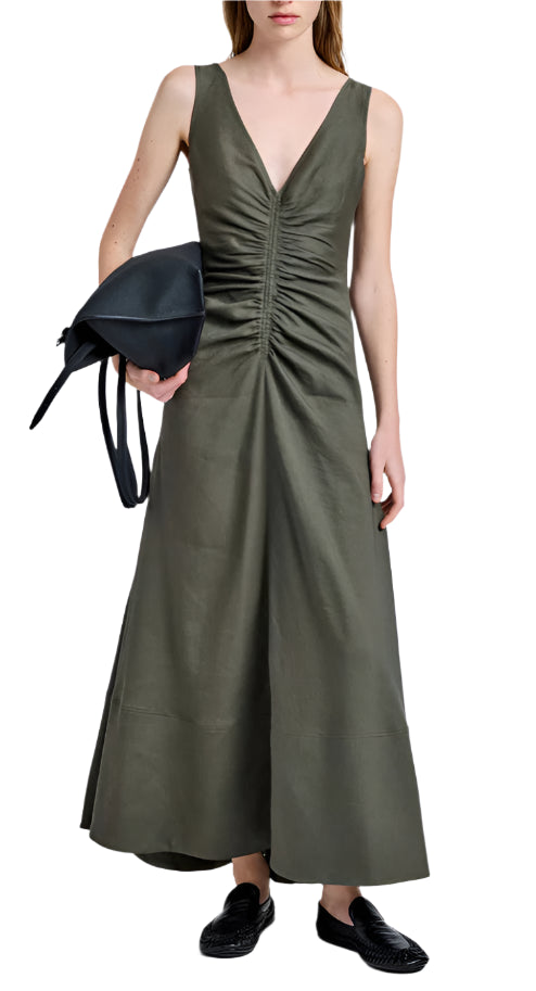 A woman wears the Remi Dress by Proenza Schouler—an olive green Eco Stretch Linen maxi with a deep V-neckline and gathered details—paired with a black bag and black loafers.