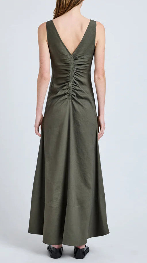 Wearing the Proenza Schouler Remi Dress in olive green eco stretch linen, featuring a sleeveless cut, V-shaped back, and gathered center detail, a person stands against a plain background.