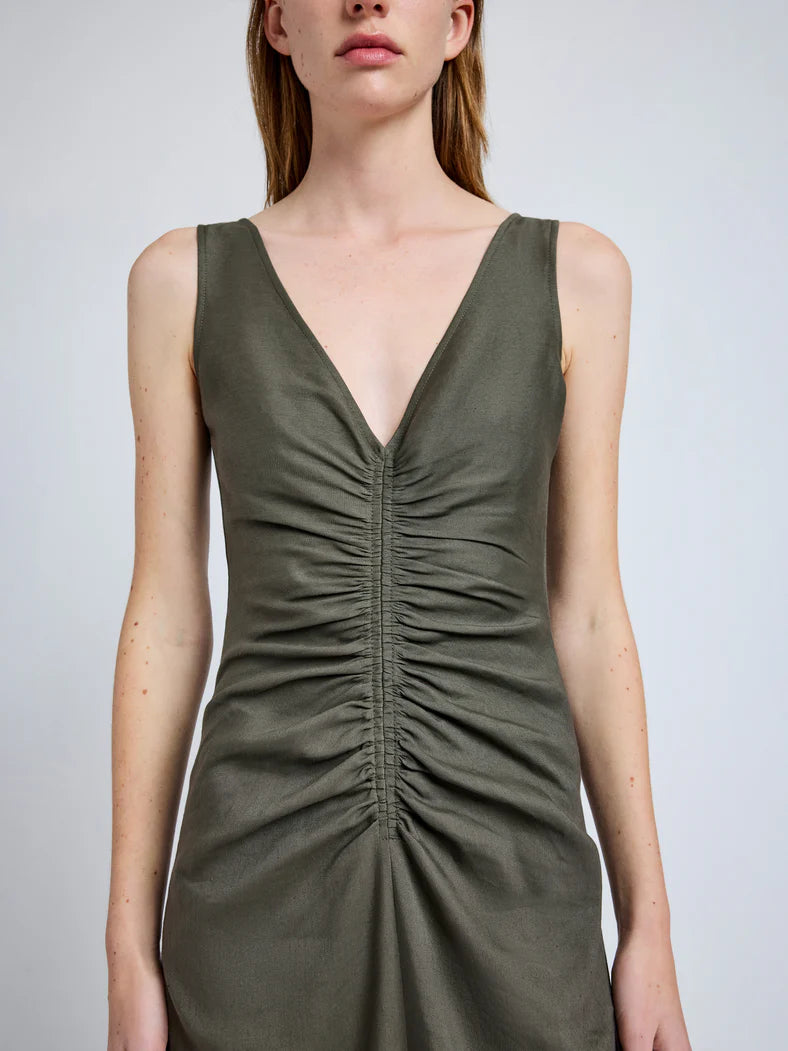 A person wears the Remi Dress by Proenza Schouler, an olive green, sleeveless design with a deep V-neckline and gathered ruching down the front, crafted from Eco Stretch Linen.