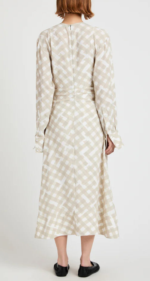 A woman faces away, wearing the Proenza Schouler Keria Printed Crepe De Chine Dress—a beige and white checkered, long-sleeved midi with a gathered twist detail—paired with black flat shoes.