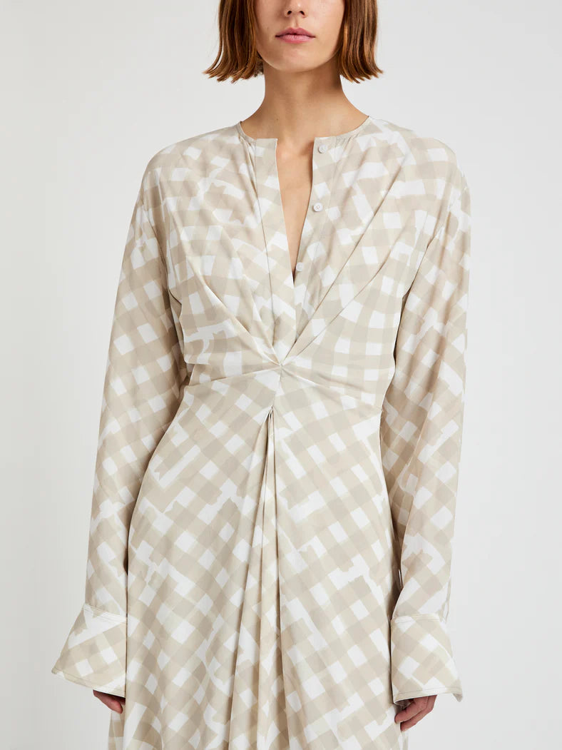 A woman wears the Proenza Schouler Keria Printed Crepe De Chine Dress in beige and white check, featuring gathered twist detail and flared cuffs, standing against a plain background.