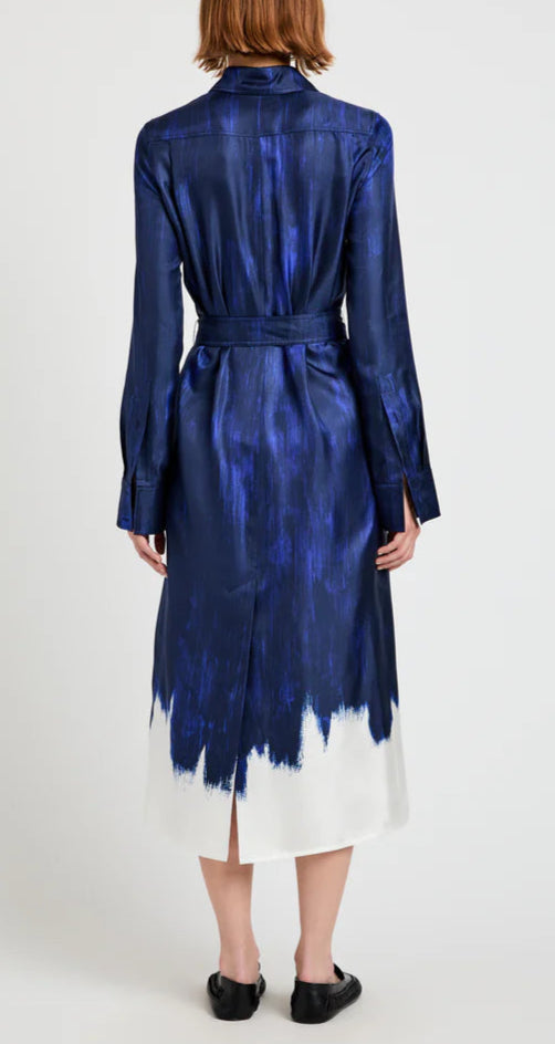 A person wearing the Proenza Schouler Amaya Silk Twill Dress, a long-sleeve blue shirtdress with a white hem and belt, stands with their back to the camera against a plain background.
