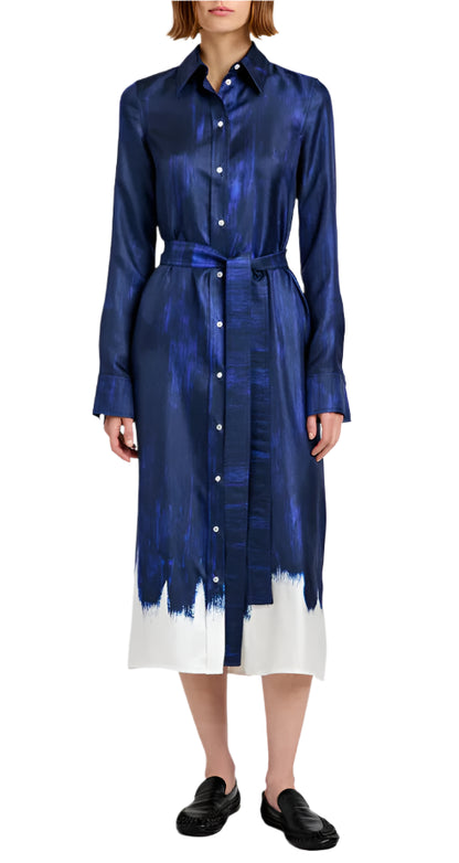 A woman wears the Proenza Schouler Amaya Silk Twill Dress—navy blue, long-sleeved with a painted white hem and matching belt—paired with black slip-on shoes.