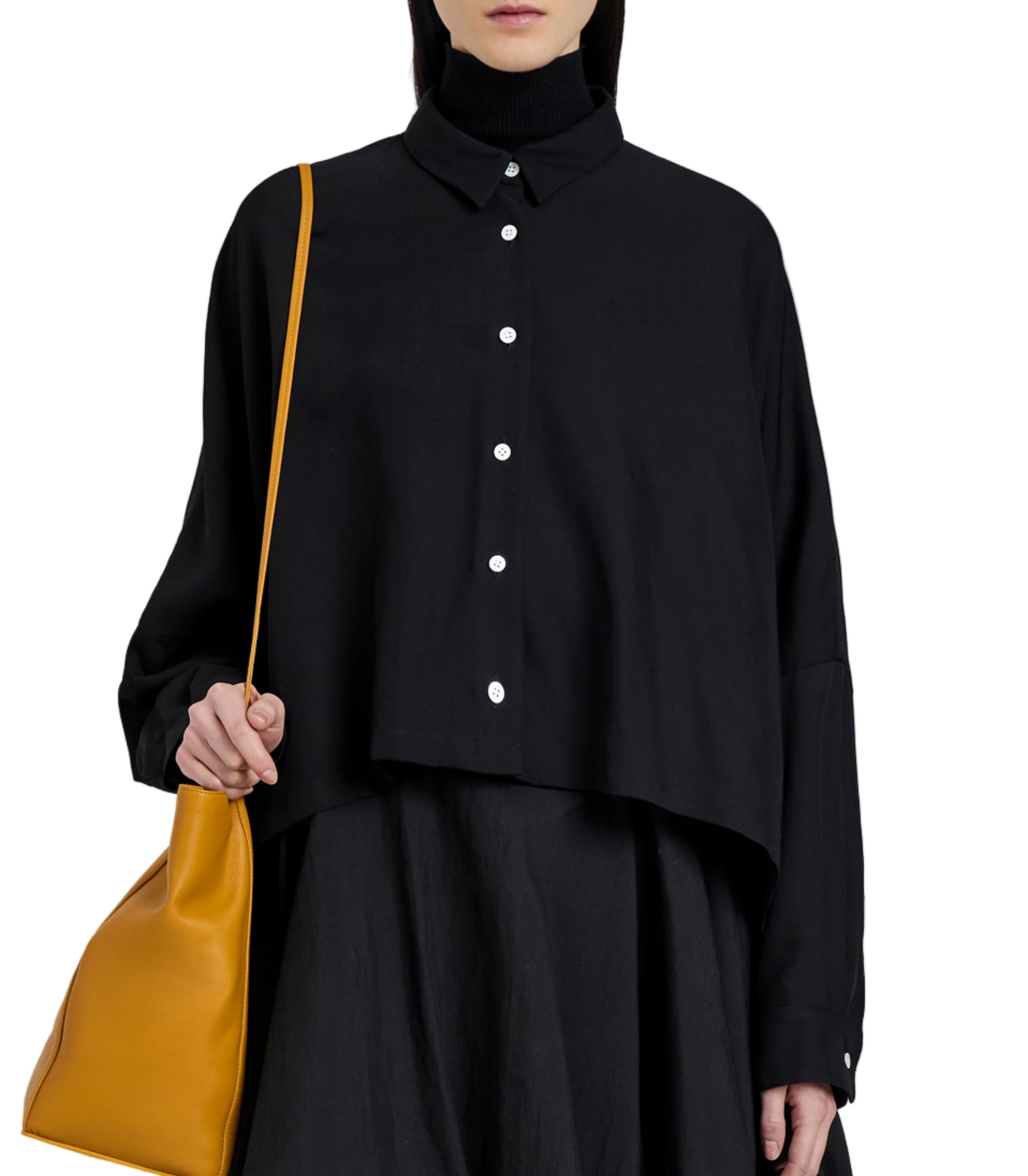 A person wearing the Zad Classic Twill Top by Proenza Schouler—a black, button-up, long-sleeve shirt with tailored details—over a black turtleneck, paired with a skirt and carrying a large mustard-yellow shoulder bag.
