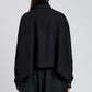 A person stands against a light backdrop, photographed from behind, wearing the Proenza Schouler Zad Classic Twill Top—a loose-fitting black jacket with a swing silhouette and cape-like back.