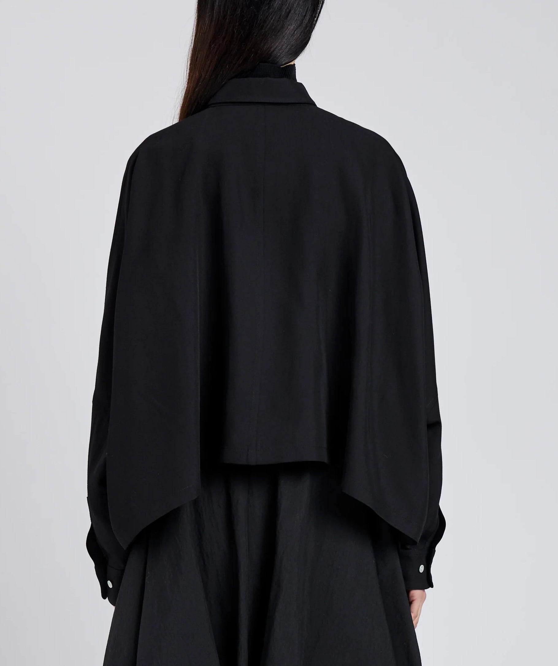 A person stands against a light backdrop, photographed from behind, wearing the Proenza Schouler Zad Classic Twill Top—a loose-fitting black jacket with a swing silhouette and cape-like back.