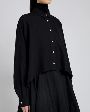 A person wears the Proenza Schouler Zad Classic Twill Top—a black collared shirt with tailored detailing and white buttons—over a black turtleneck and skirt, shown waist up against a plain backdrop.