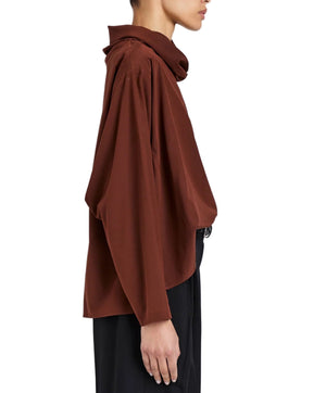 A person wearing the Proenza Schouler Solene Silk Viscose Top—loose, long-sleeved with a funnel neck—and black pants, shown in profile against a plain background.