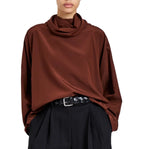 A person wearing the Proenza Schouler Solene Silk Viscose Top in brown with long sleeves and a funnel neck, tucked into high-waisted black pleated pants with a black woven belt.