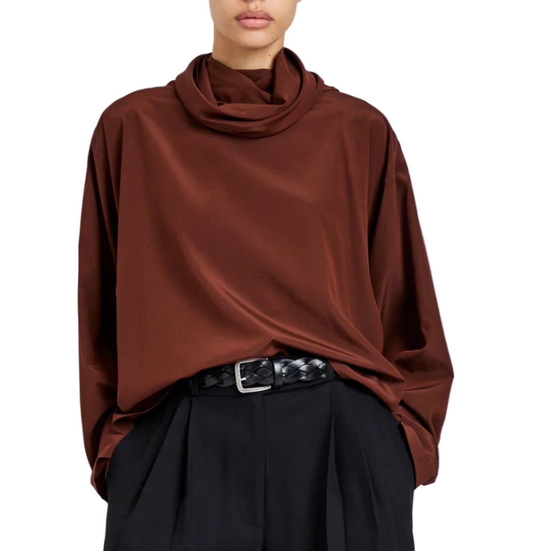 A person wearing the Proenza Schouler Solene Silk Viscose Top in brown with long sleeves and a funnel neck, tucked into high-waisted black pleated pants with a black woven belt.