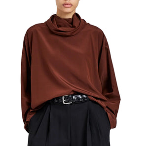 A person wearing the Proenza Schouler Solene Silk Viscose Top in brown with long sleeves and a funnel neck, tucked into high-waisted black pleated pants with a black woven belt.