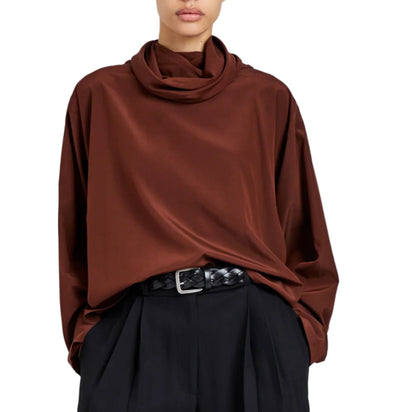 A person wearing the Proenza Schouler Solene Silk Viscose Top in brown with long sleeves and a funnel neck, tucked into high-waisted black pleated pants with a black woven belt.