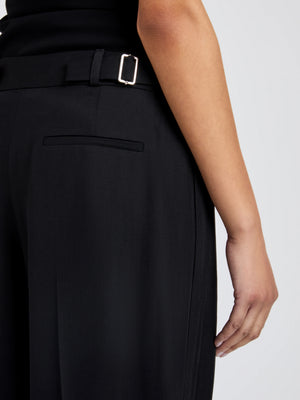 A person wears the Proenza Schouler Patricia Classic Twill Pant in black, featuring wide legs and a side buckle detail, shown from the waist down with their right arm at their side.