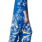 A woman wearing the Figue Paolina Kaftan in blue, featuring white floral patterns and a tassel-trimmed hemline, paired with silver sandals, stands sideways against a white background.