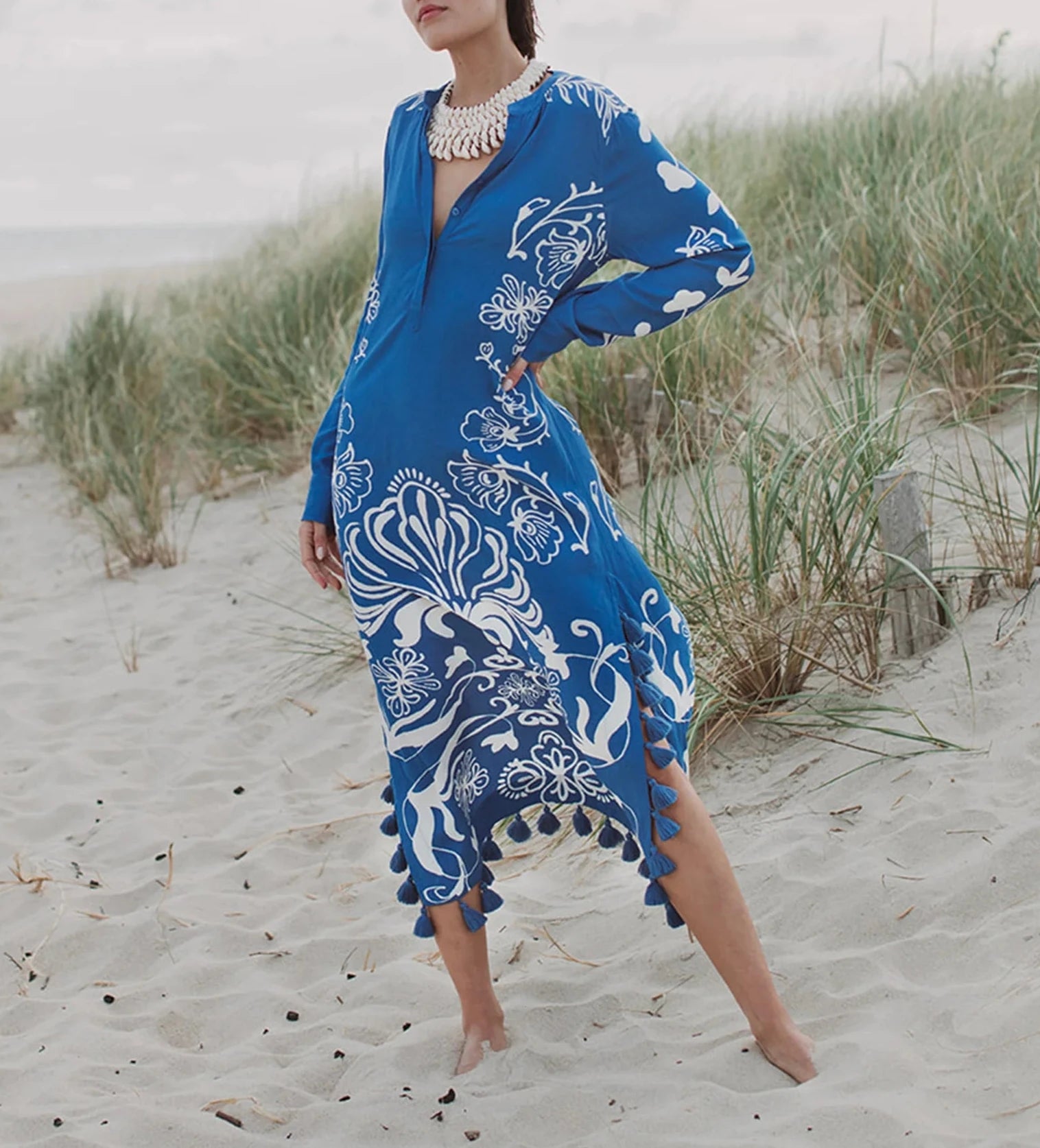 A person stands on a sandy beach with grass dunes in the background, wearing the elegant Figue Paolina Kaftan, which features a blue and white patterned design with a split neckline and tassel trimmed hemline.