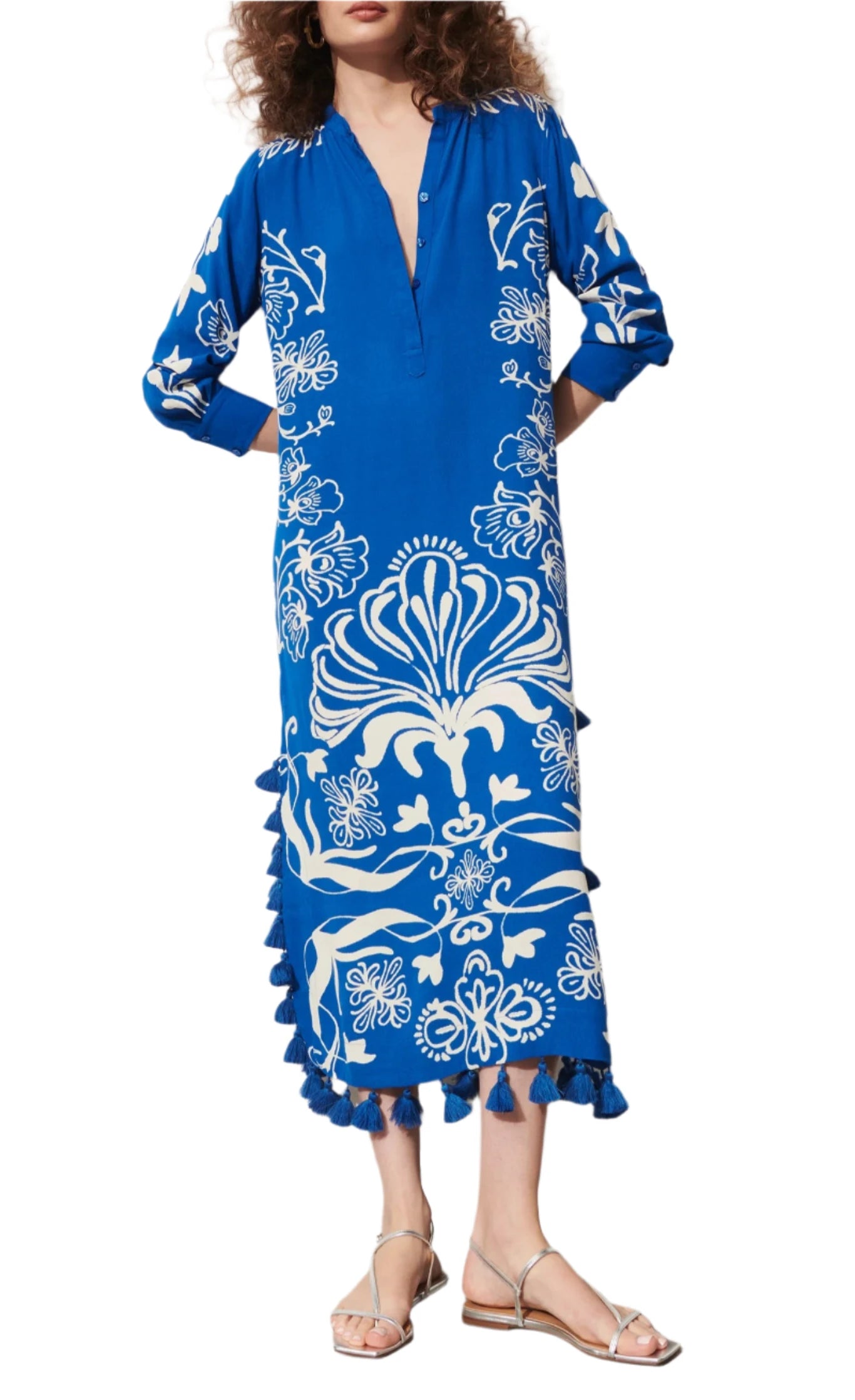 A person wearing the Figue Paolina Kaftan featuring a blue and white floral pattern, a split neckline, long sleeves, and tassel details along the hemline, paired with silver sandals.