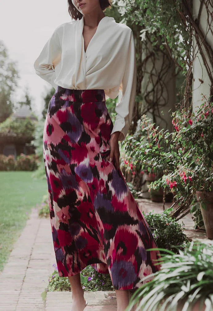 A person wearing a white blouse and the Figue Isla Skirt, a long, multicolored circle cut skirt with a trapunto waistband from Figue, stands outdoors near green foliage.