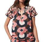 A person wearing a Figue Rosie Top by Figue, featuring a V-neckline and lace details at the shoulders. The short-sleeved, patterned blouse showcases pink, brown, and black abstract shapes and includes custom embroidery for an extra touch of elegance.