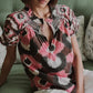 A person wearing the Rosie Top by Figue, featuring custom embroidery and lace detailing, is seated on a green couch with floral pillows.