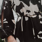 A person wearing a black and white abstract patterned Figue Kasey Tunic, which features a boat neckline, long sleeves, and a thin belt, stands outdoors.