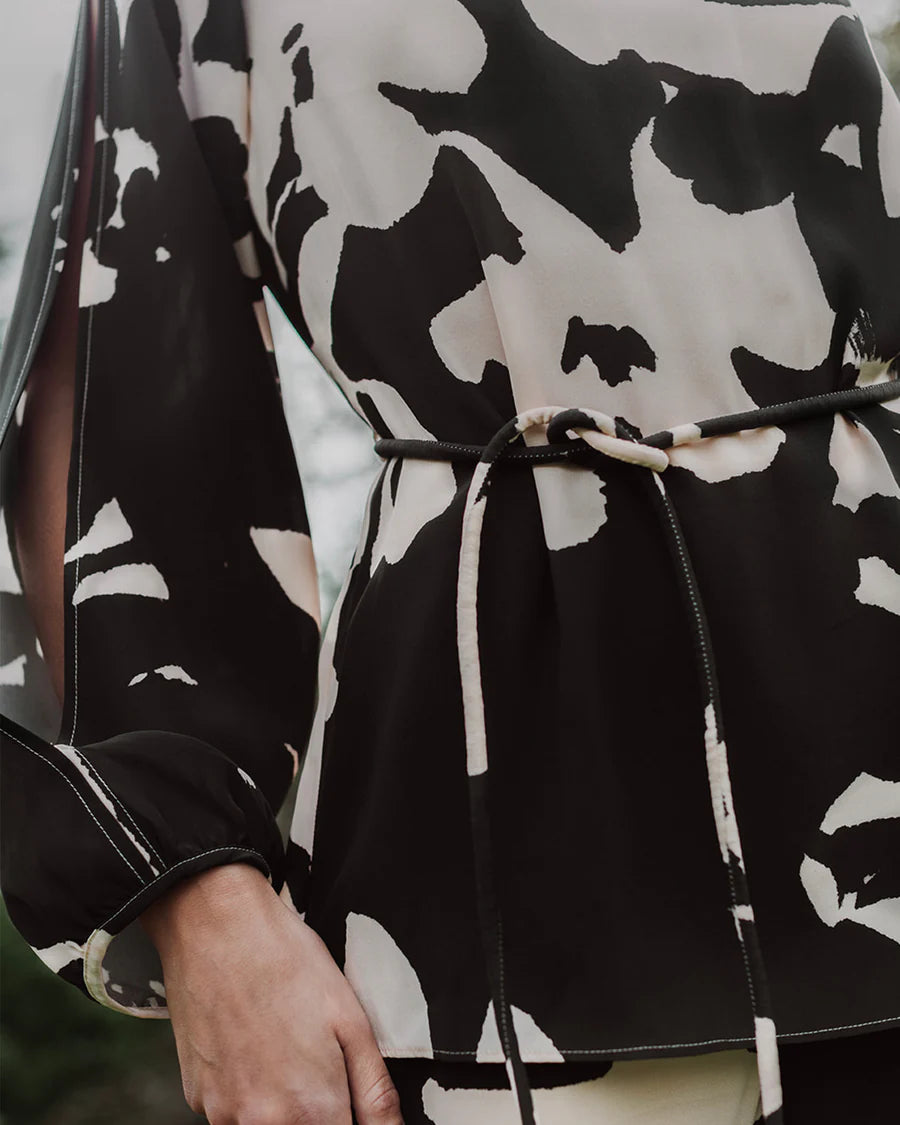 Close-up of a person wearing the elegant Figue Kasey Tunic, featuring a black and white patterned top, long sleeves, and a thin belt accentuating the waist. The boat neckline adds an extra touch of sophistication to the outfit.