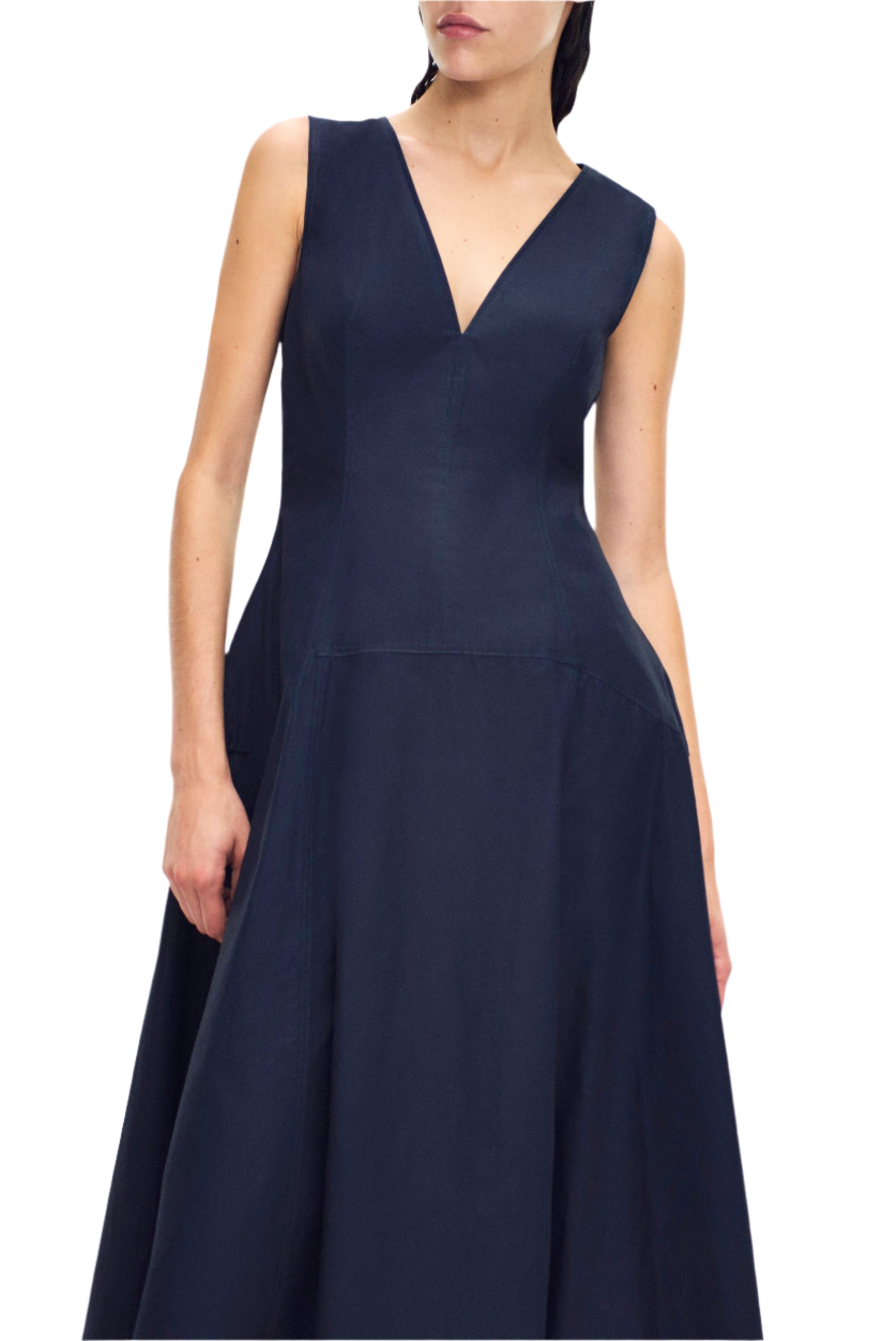 Clad in a sleeveless, navy blue Adam Lippes Harlow Dress made from soft cotton poplin, an individual stands elegantly against a plain backdrop.