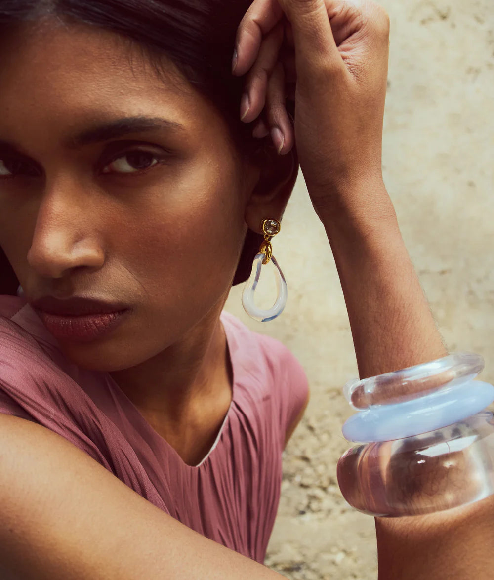 A woman wears a pink top, the Lizzie Fortunato Ridge Cuff, and gold and clear teardrop earrings as she poses with her hand near her face against a neutral background.