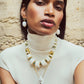 A woman in a white sleeveless turtleneck wears bold white and gold jewelry, including Lizzie Fortunato's La Paz Earrings, with porcelain bead drops, and poses against a textured stone wall.