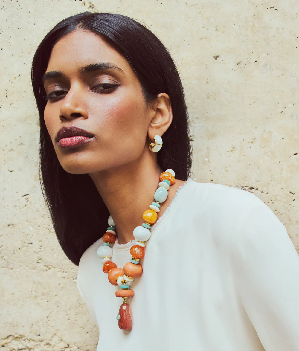 Woman with long black hair wears a white top, colorful handcrafted beaded necklace, and Lizzie Fortunato Zellige Hoops, standing against a textured beige wall.
