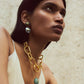 A woman with straight dark hair wears a white top, a large gold chain necklace with a blue pendant, and Lizzie Fortunato's Dorothy Earrings—gold and blue statement pieces featuring green jade—against a neutral textured background.