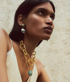 A woman with straight dark hair wears a white top, a large gold chain necklace with a blue pendant, and Lizzie Fortunato's Dorothy Earrings—gold and blue statement pieces featuring green jade—against a neutral textured background.