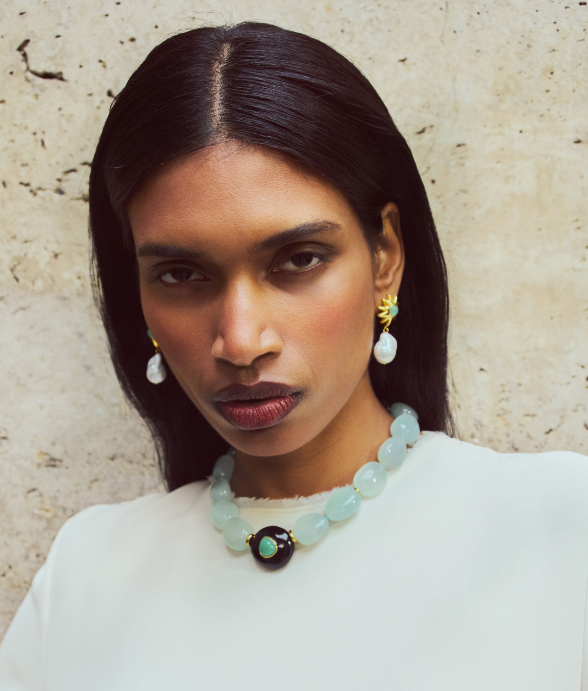 A woman with long dark hair, pearl earrings, and a white top wears the Lizzie Fortunato Andalusia Collar—a blue beaded necklace with turquoise accents and a black pendant—against a textured beige wall.