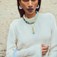 A woman in a cream long-sleeve top stands against a textured wall, wearing layered turquoise beaded necklaces, gold jewelry, and Lizzie Fortunato's Organic Gold Hoops.