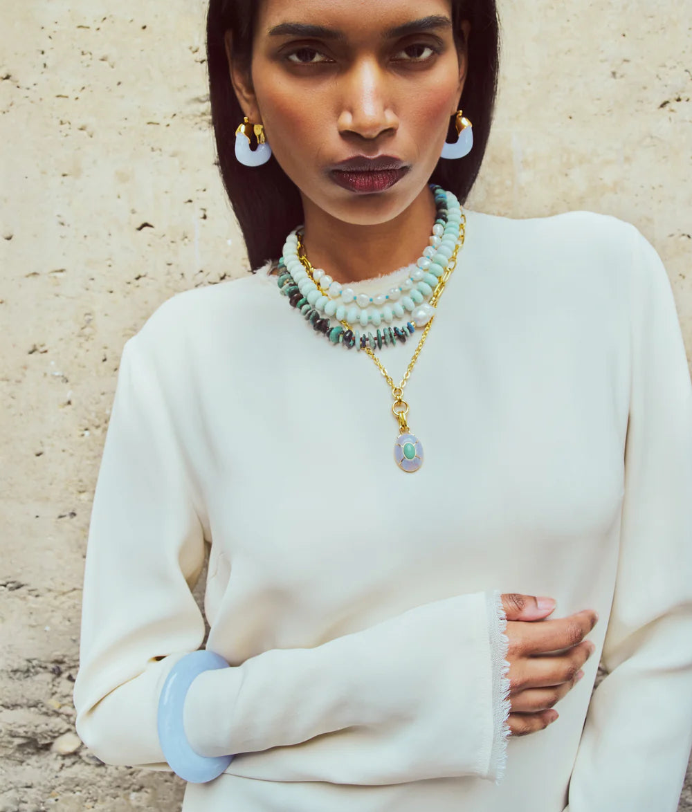 A woman in a cream long-sleeve top stands against a textured wall, wearing layered turquoise beaded necklaces, gold jewelry, and Lizzie Fortunato's Organic Gold Hoops.