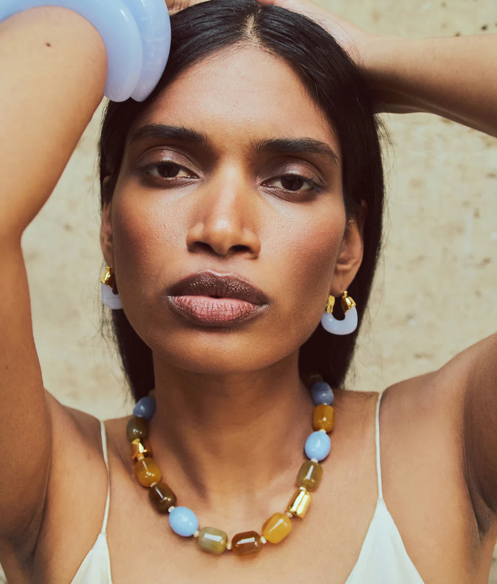 A woman with long dark hair poses with her arms raised, wearing Lizzie Fortunato’s Organic Mini Gold Hoops and pastel jewelry against a neutral background.