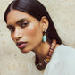 A person with straight dark hair wears a white top and the Las Palmas Collar by Lizzie Fortunato, along with dangling earrings featuring turquoise and black beads, against a textured light backdrop.