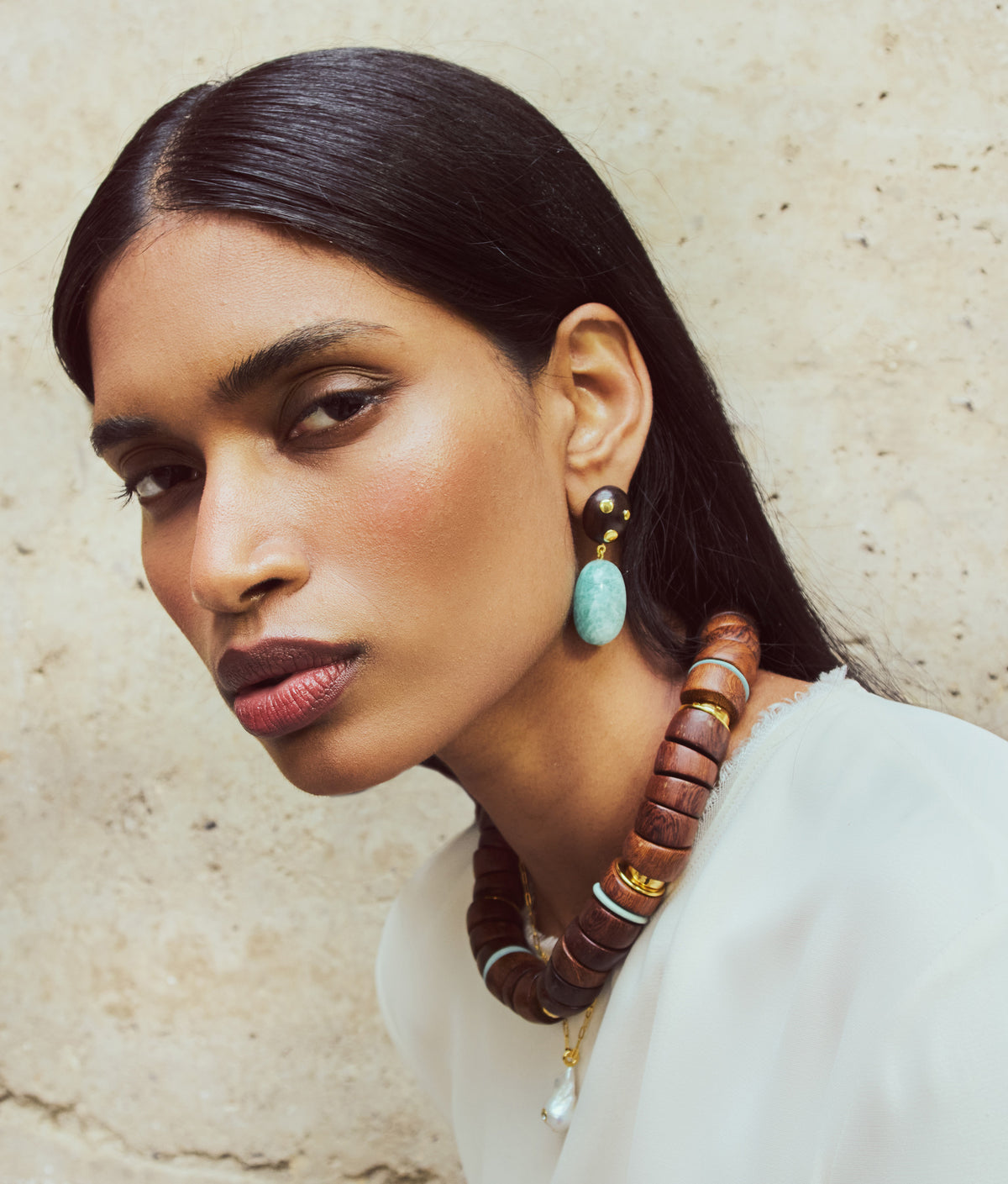 A person with straight dark hair wears a white top and the Las Palmas Collar by Lizzie Fortunato, along with dangling earrings featuring turquoise and black beads, against a textured light backdrop.