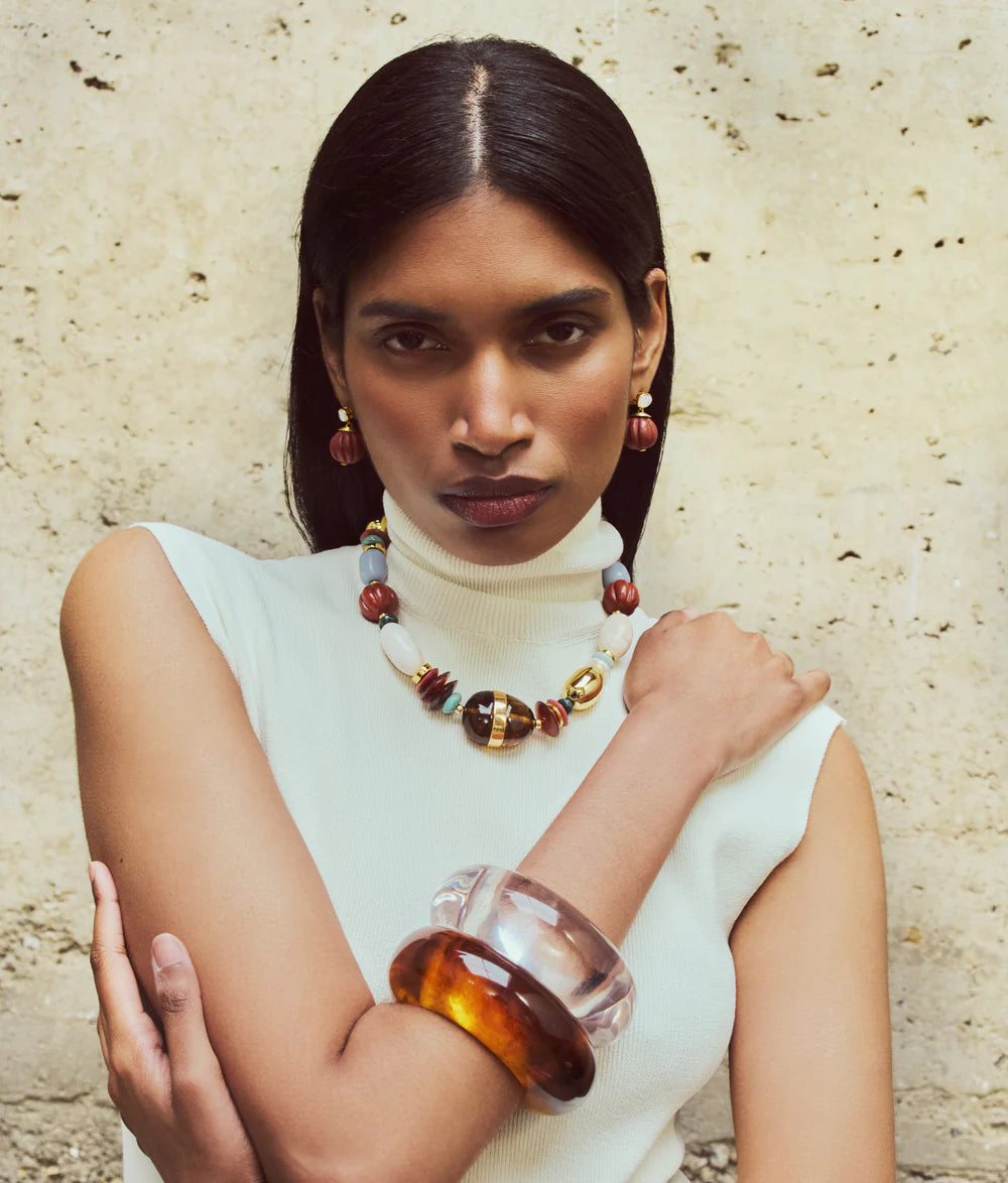 A woman with straight dark hair wears a sleeveless white turtleneck, the Lizzie Fortunato Kona Collar necklace, matching earrings, and large amber and clear bangles, standing against a textured wall.