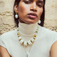 A person wearing a white sleeveless turtleneck, chunky white and gold necklace, matching earrings, and the Lizzie Fortunato La Paz Pendant Necklace poses against a textured beige wall.