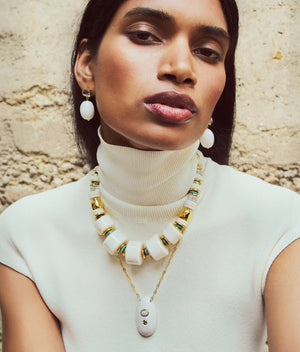 A person wearing a white sleeveless turtleneck, chunky white and gold necklace, matching earrings, and the Lizzie Fortunato La Paz Pendant Necklace poses against a textured beige wall.