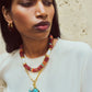 A woman in a white top and gold hoops stands against a textured beige wall, wearing the Lizzie Fortunato Floria Heart Necklace featuring layered beaded strands, a turquoise heart pendant, and a gold-plated chain.
