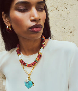 A woman in a white top and gold hoops stands against a textured beige wall, wearing the Lizzie Fortunato Floria Heart Necklace featuring layered beaded strands, a turquoise heart pendant, and a gold-plated chain.