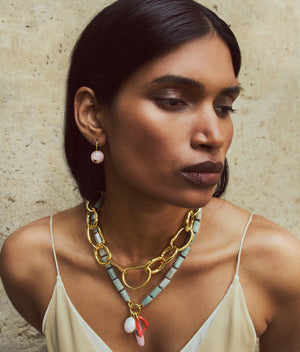 A woman with straight dark hair wears a cream top, the Lizzie Fortunato Cortez Necklace with green and pink accents, and matching pink opal drop earrings against a textured beige background.