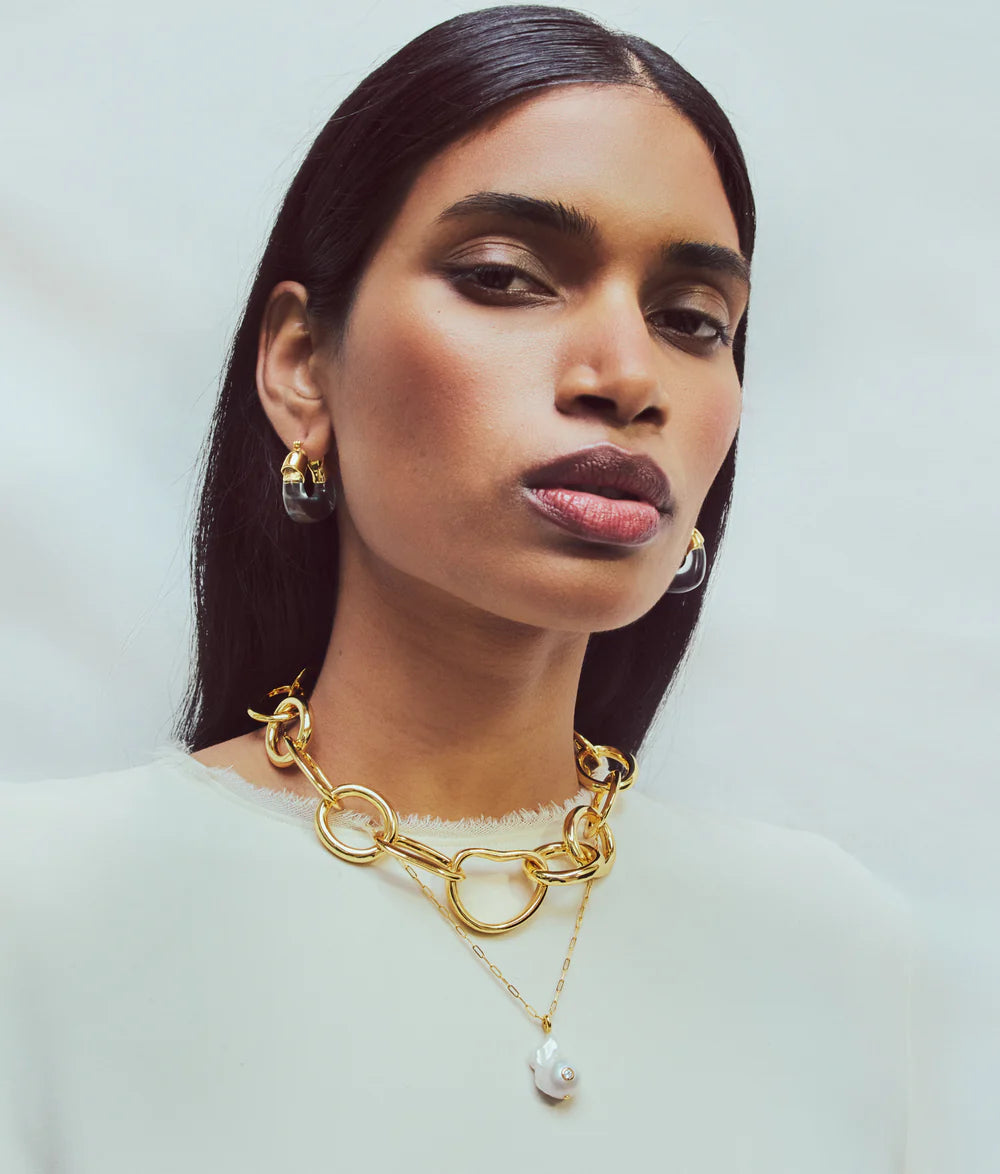 A woman with straight dark hair wears gold hoop earrings and the Lizzie Fortunato Oasis Necklace, a delicate gold chain with a pearl pendant, against a plain light background.