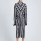 A person with short hair, wearing white shoes, stands facing away in the Proenza Schouler Amira Dress—a knee-length, long-sleeved shirtdress made from printed silk habotai with black, white, and blue stripes in vertical and chevron patterns.