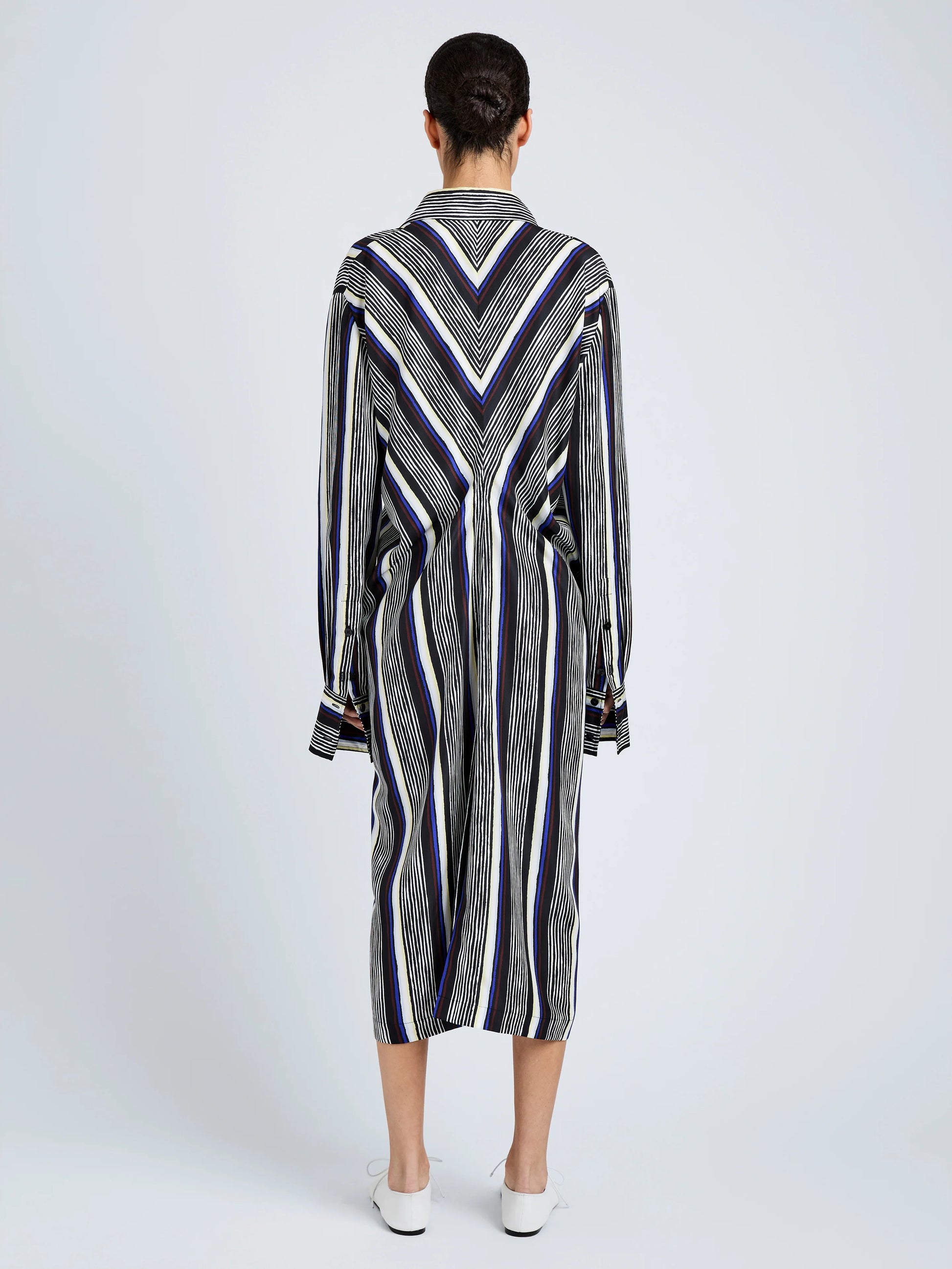 A person with short hair, wearing white shoes, stands facing away in the Proenza Schouler Amira Dress—a knee-length, long-sleeved shirtdress made from printed silk habotai with black, white, and blue stripes in vertical and chevron patterns.