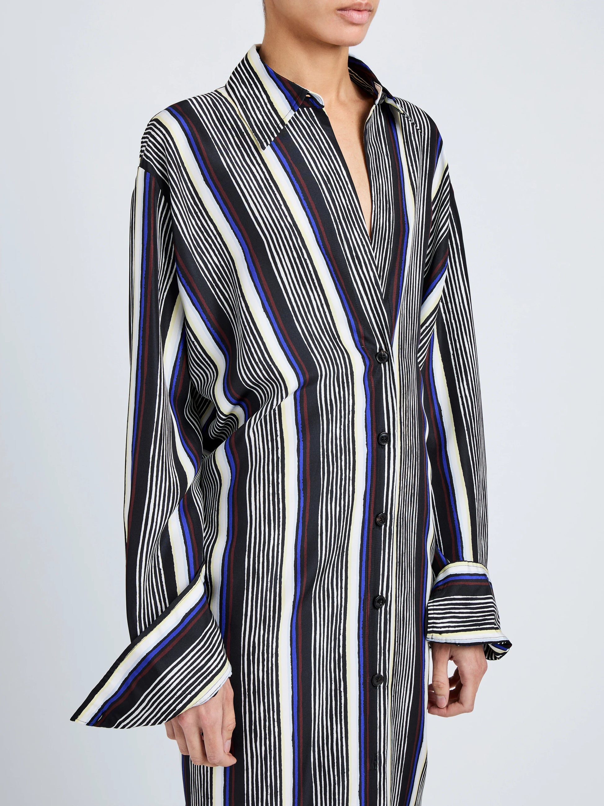 A person wears the Amira Dress by Proenza Schouler, a long-sleeved, button-down shirtdress with vertical black, white, and blue stripes in luxe Printed Silk Habotai, standing against a plain background.